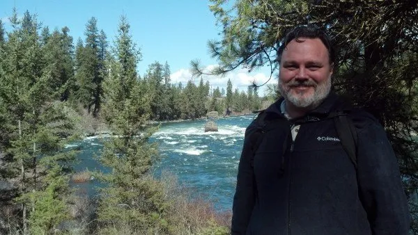 A man with a beard stands in front of a river surrounded by dense pine trees, wearing a black jacket and carrying a backpack.