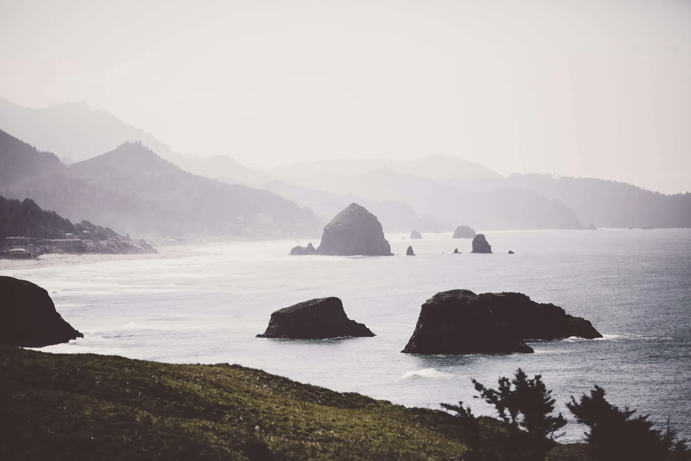 A coastal scene with sea stacks emerging from the ocean, lightly misted mountains in the background, and hilly greenery in the foreground.