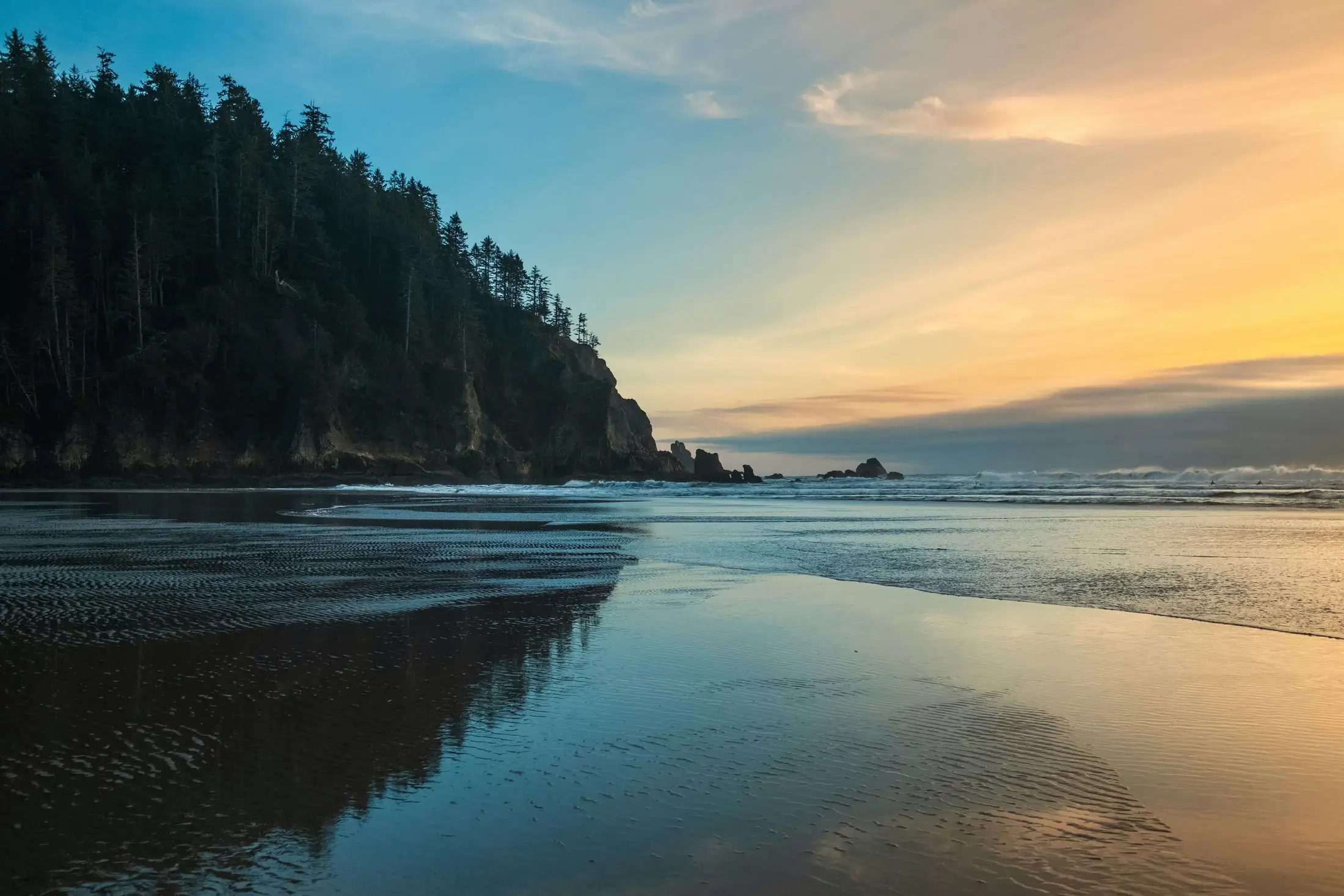A serene beach with calm waves, a reflective shoreline, and a forested cliff under a sunset sky.