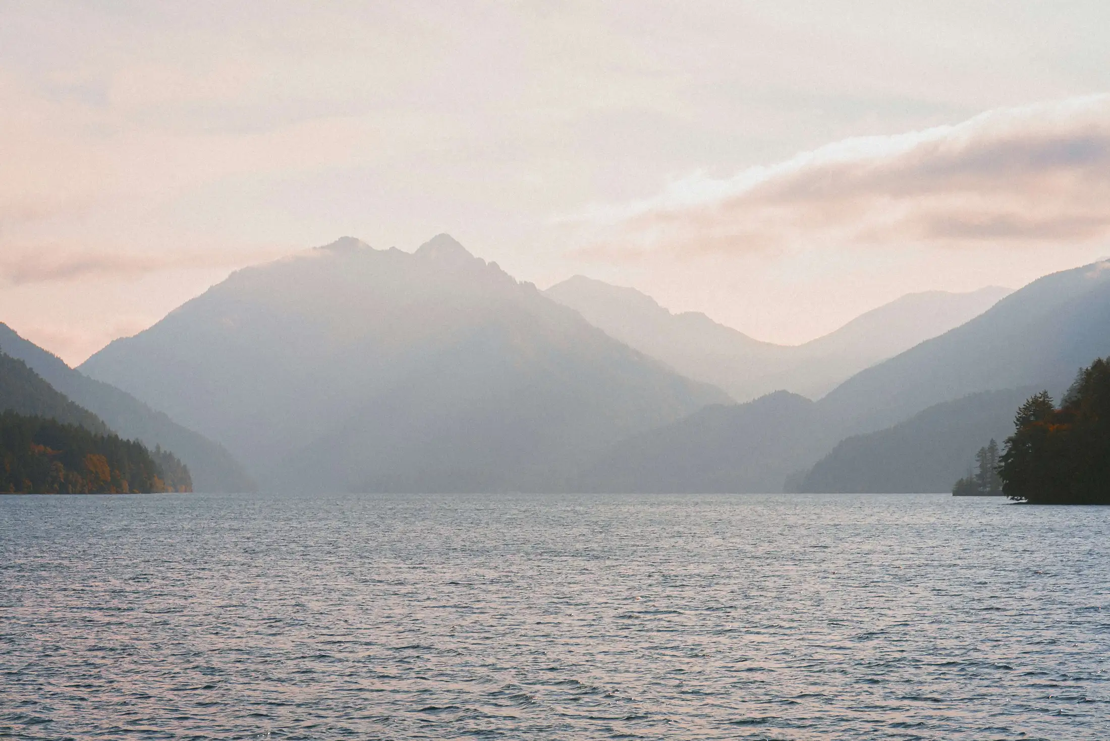 A serene lake with distant mountains partially shrouded in mist and backlit by the soft glow of a setting or rising sun.