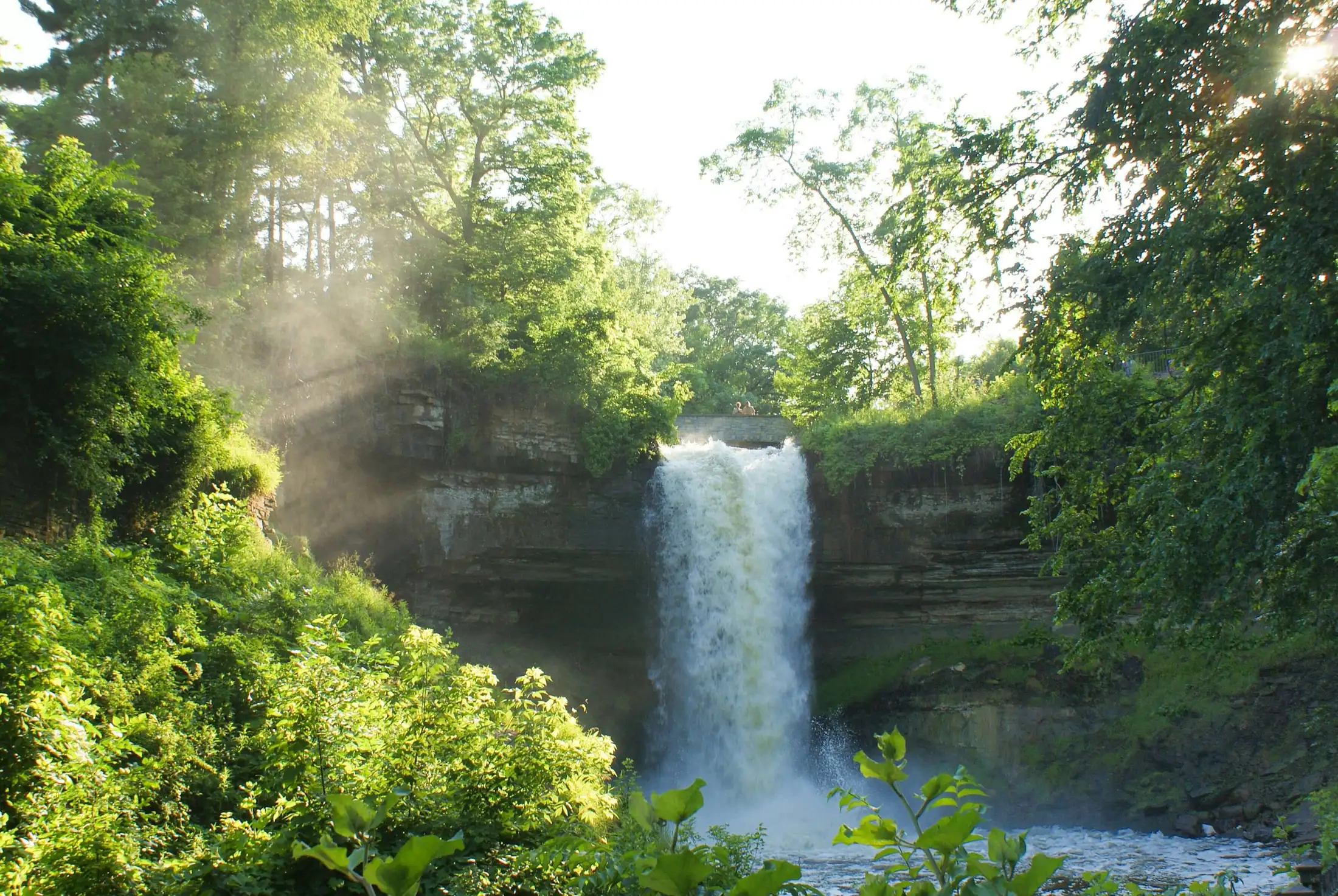 A waterfall cascades over a rocky ledge surrounded by lush greenery with mist rising in the sunlight.