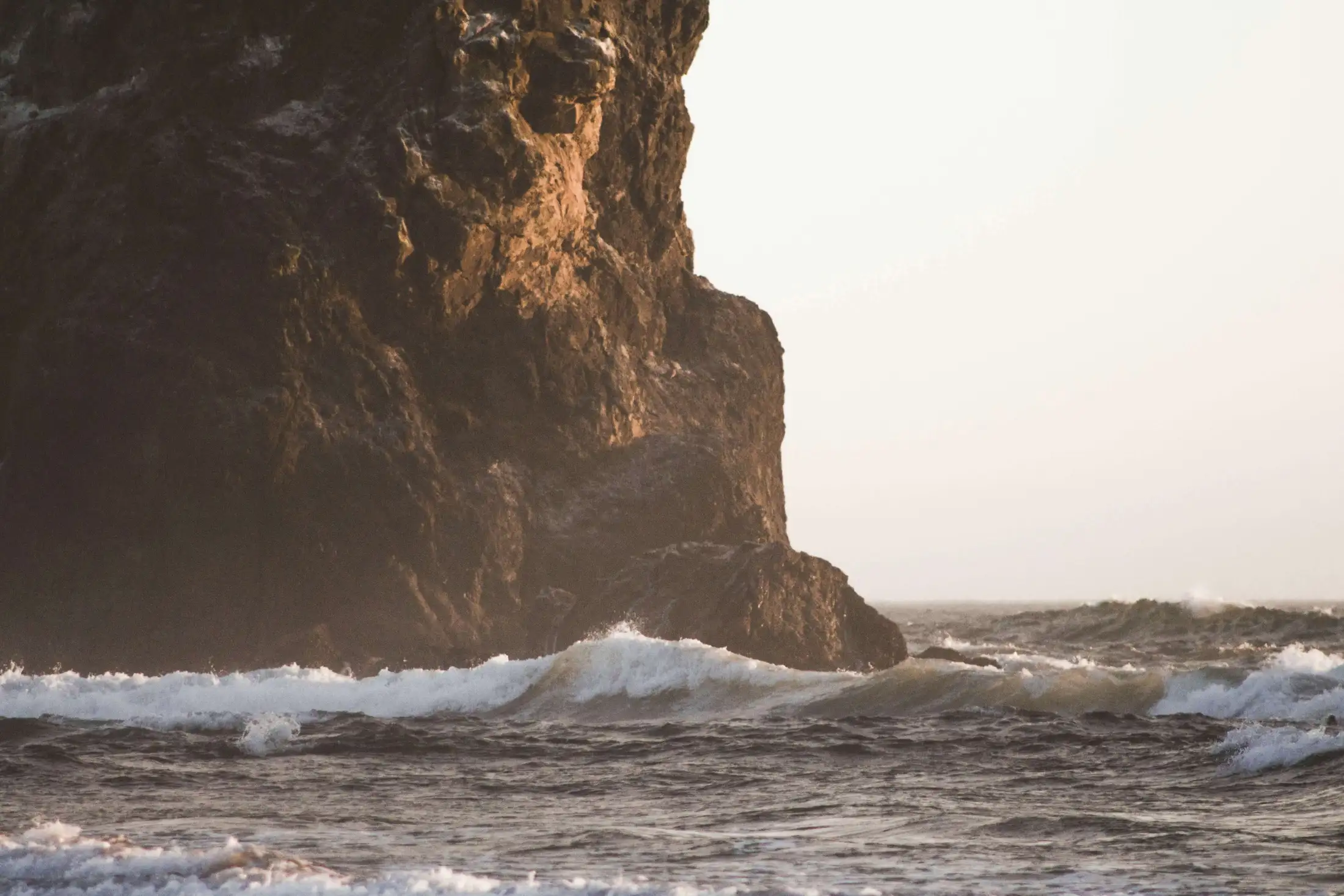 Large rocky cliff face next to ocean waves at sunset.