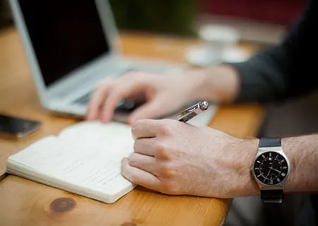 A person writes in a notebook with a pen while working on a laptop.