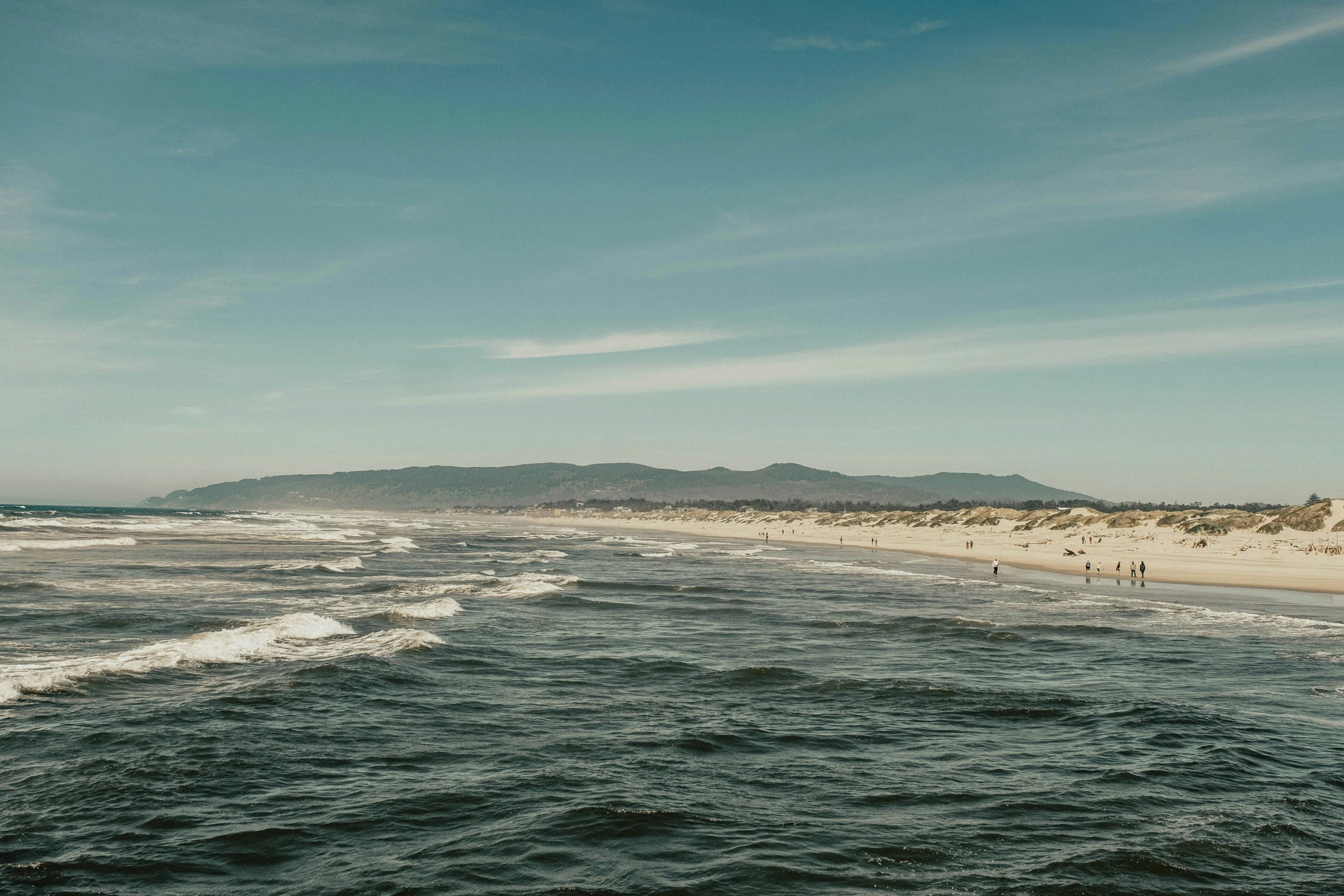 A wide beach with gentle waves and sparse clouds. Several people walk along the sandy shore with a distant range of hills in the background.