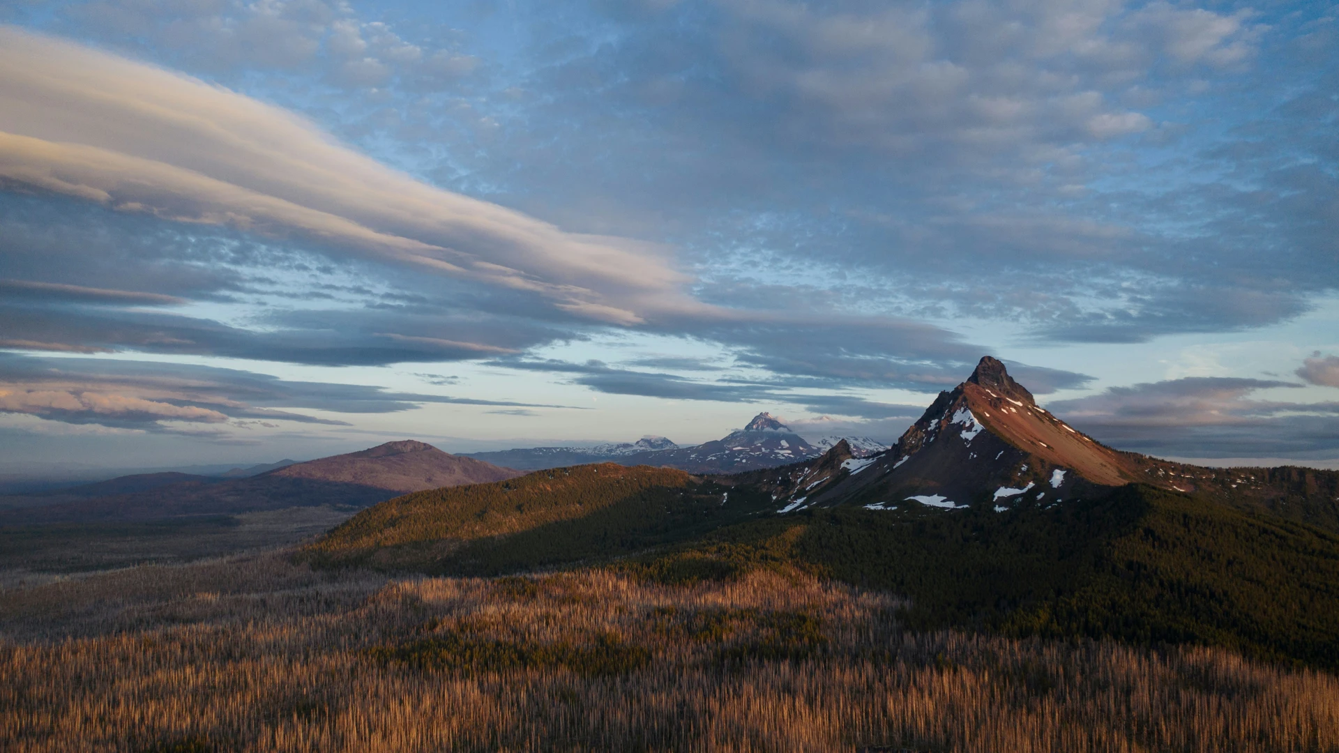 A rugged mountain landscape under a partly cloudy sky, with sparse snow on the peaks and expansive, forested terrain.