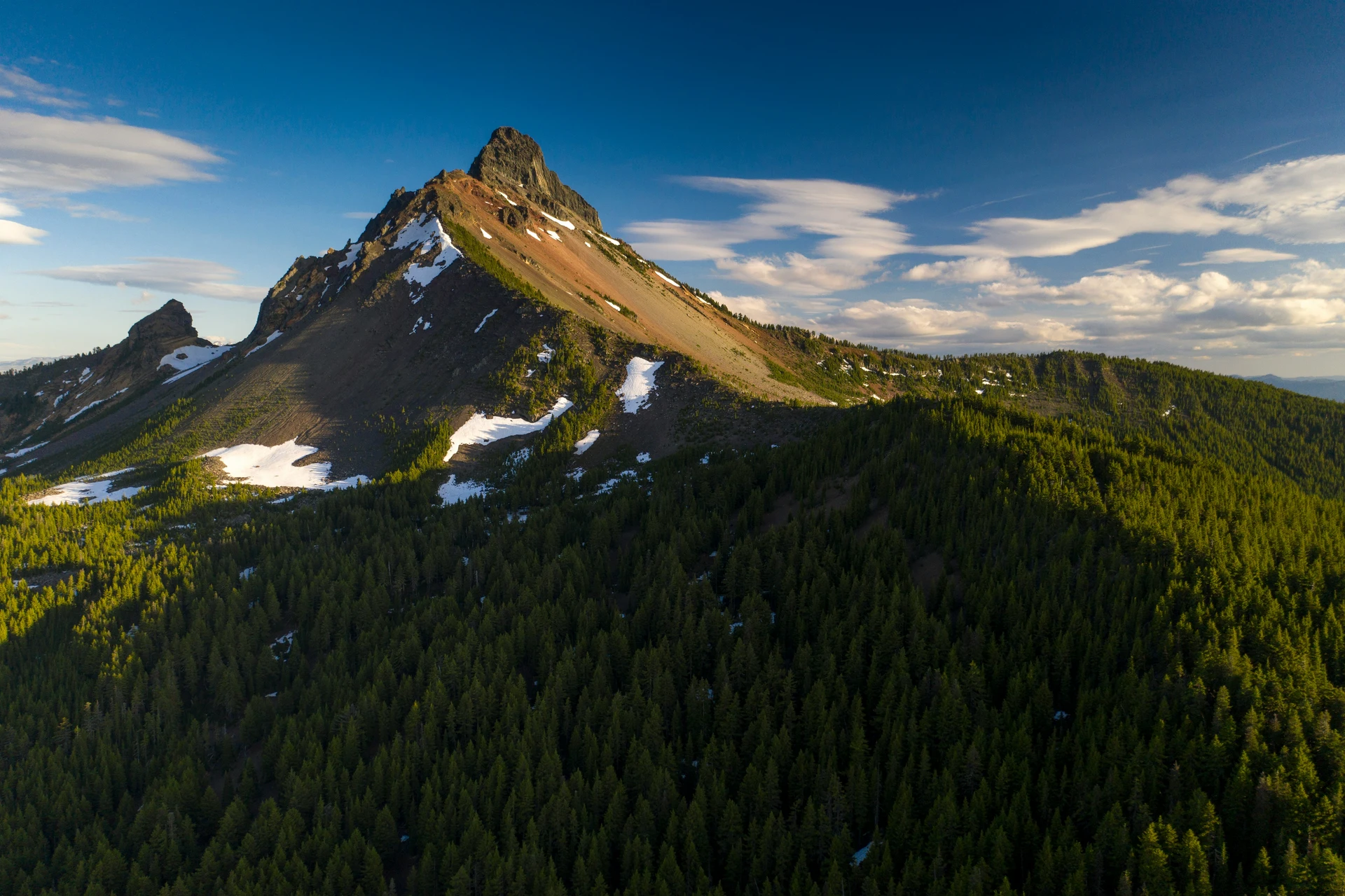 Aerial view of a mountain peak with patches of snow, surrounded by dense green forest under a blue sky with scattered clouds.