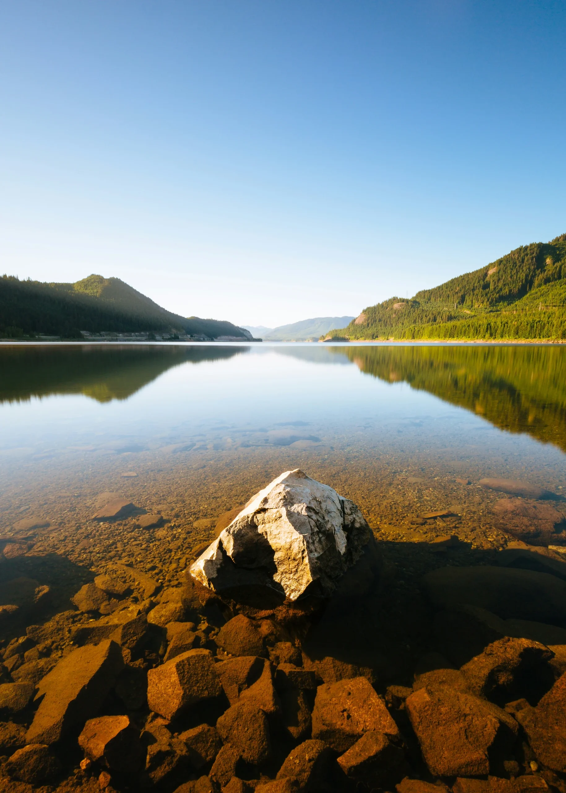 A large rock sits in shallow, clear water near the shore of a calm lake surrounded by mountains under a bright blue sky.
