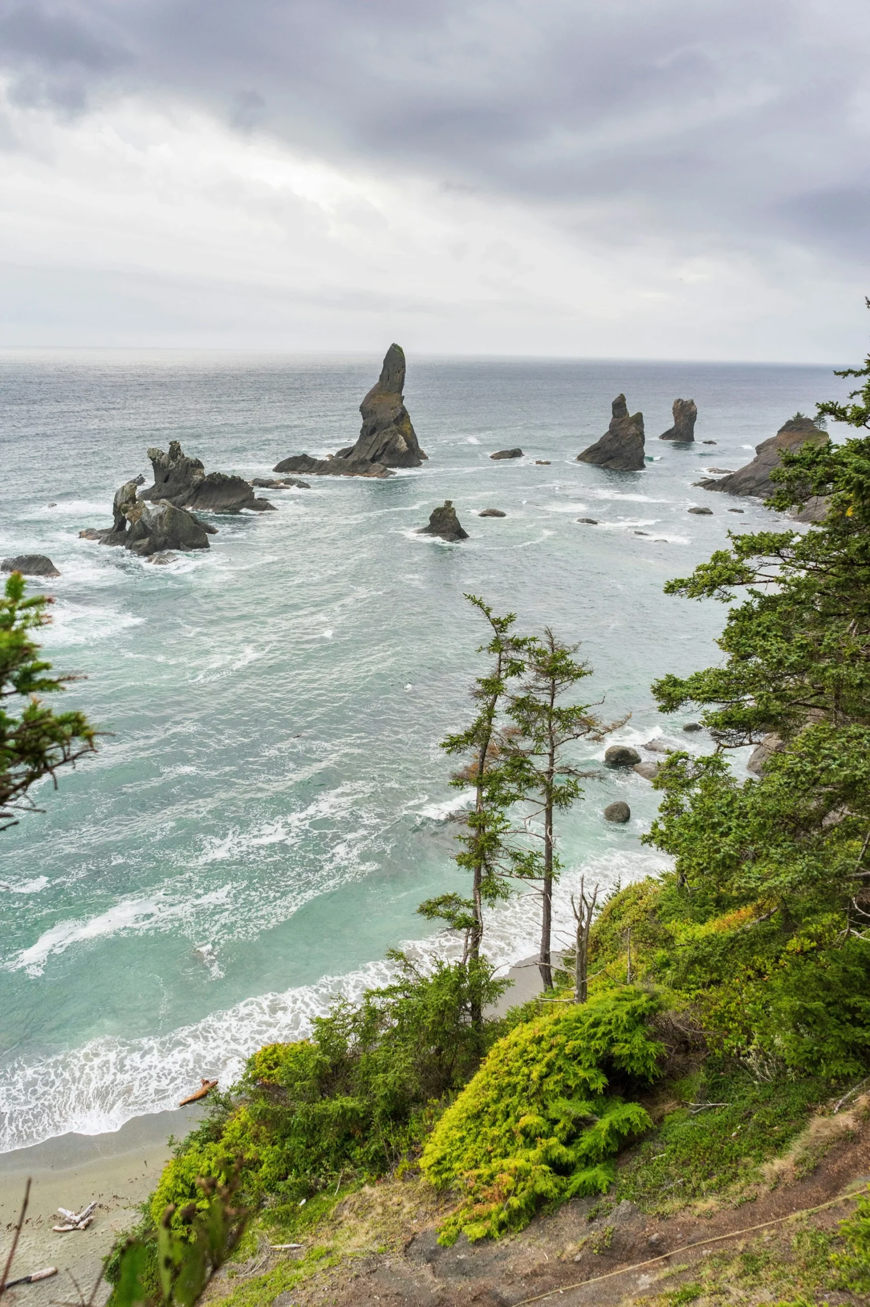 Rocky sea stacks in the Pacific Ocean near a lush, green coastline under a cloudy sky.