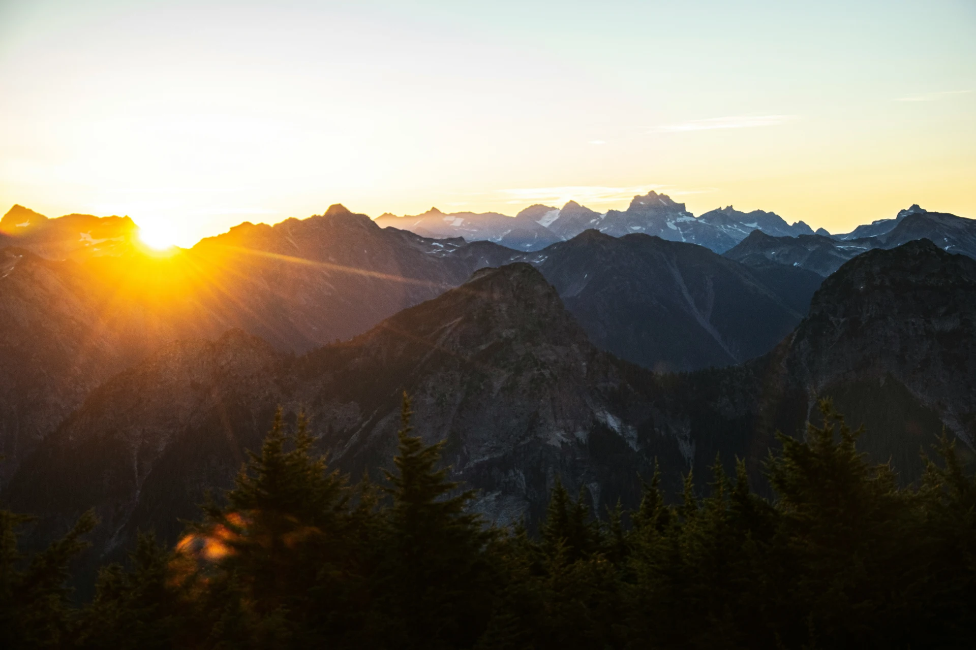 Sun rising over a mountainous landscape with trees in the foreground, casting a golden light on the peaks.