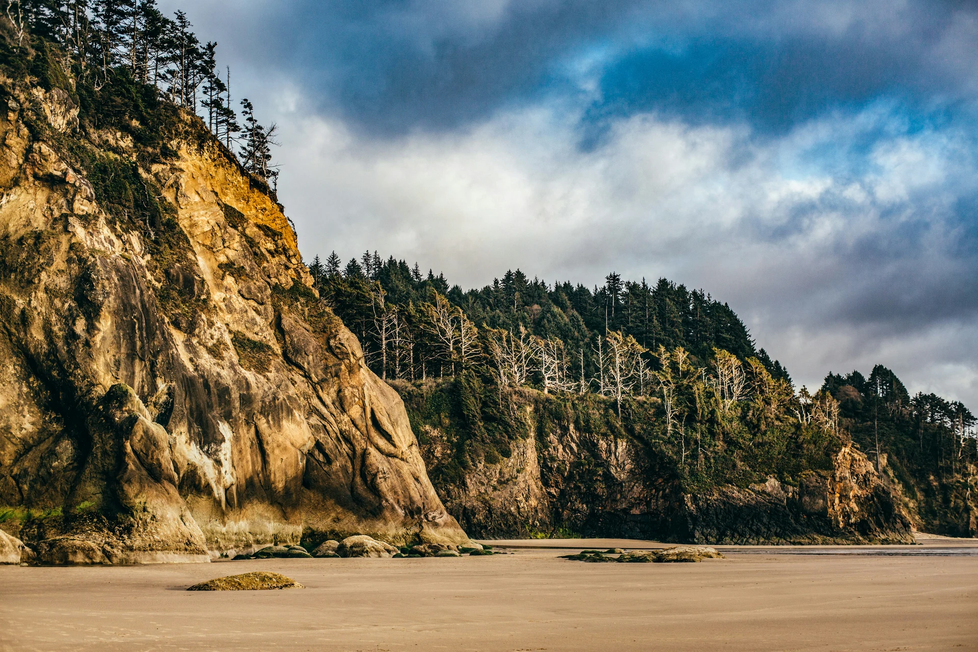 A rocky, tree-covered cliff borders a sandy beach under a cloudy sky. The beach is expansive, with a few large rocks scattered around. Dense forest covers much of the cliffside.