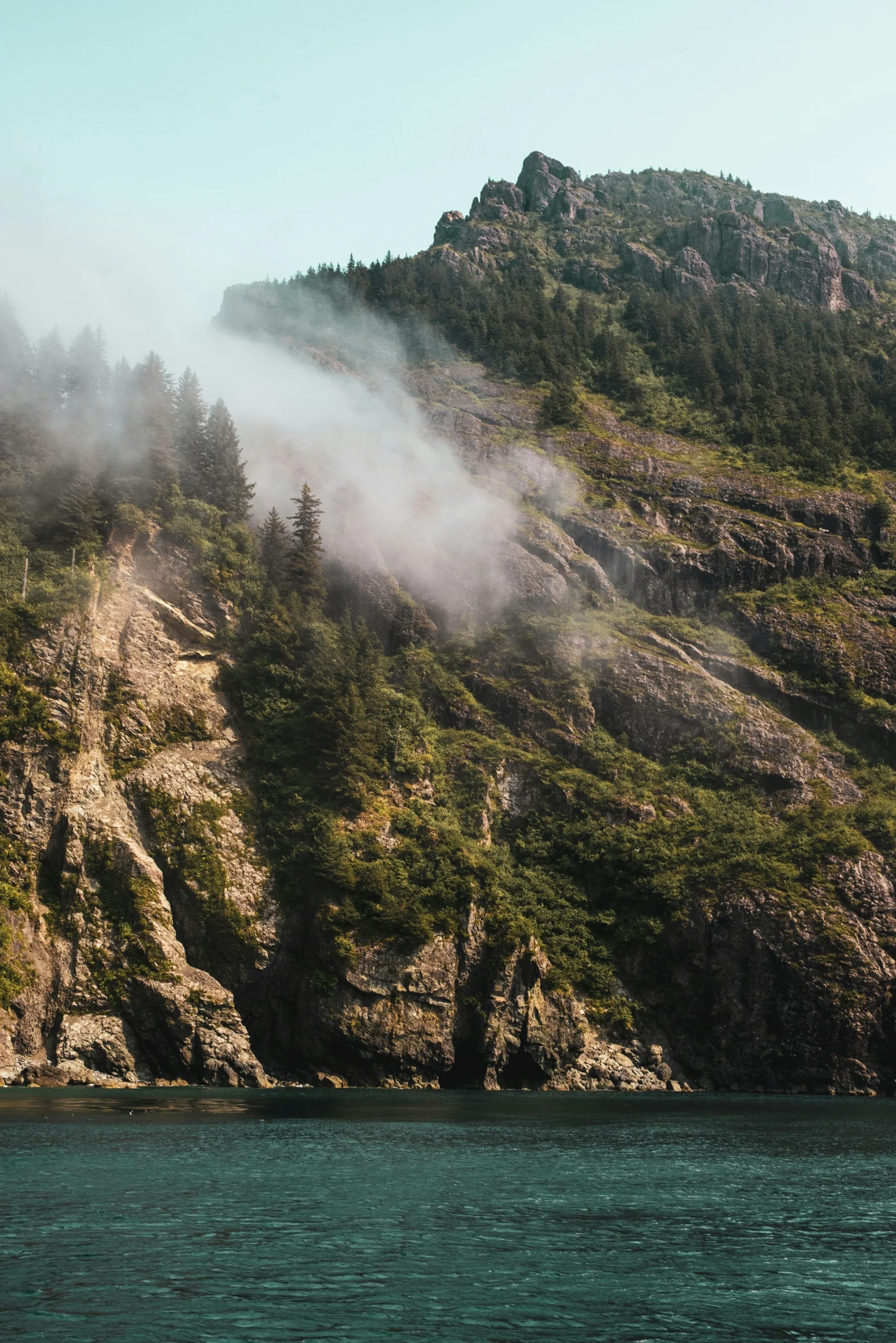 A steep, rocky cliff covered with pine trees and enveloped in fog rises from a turquoise body of water against a clear sky.