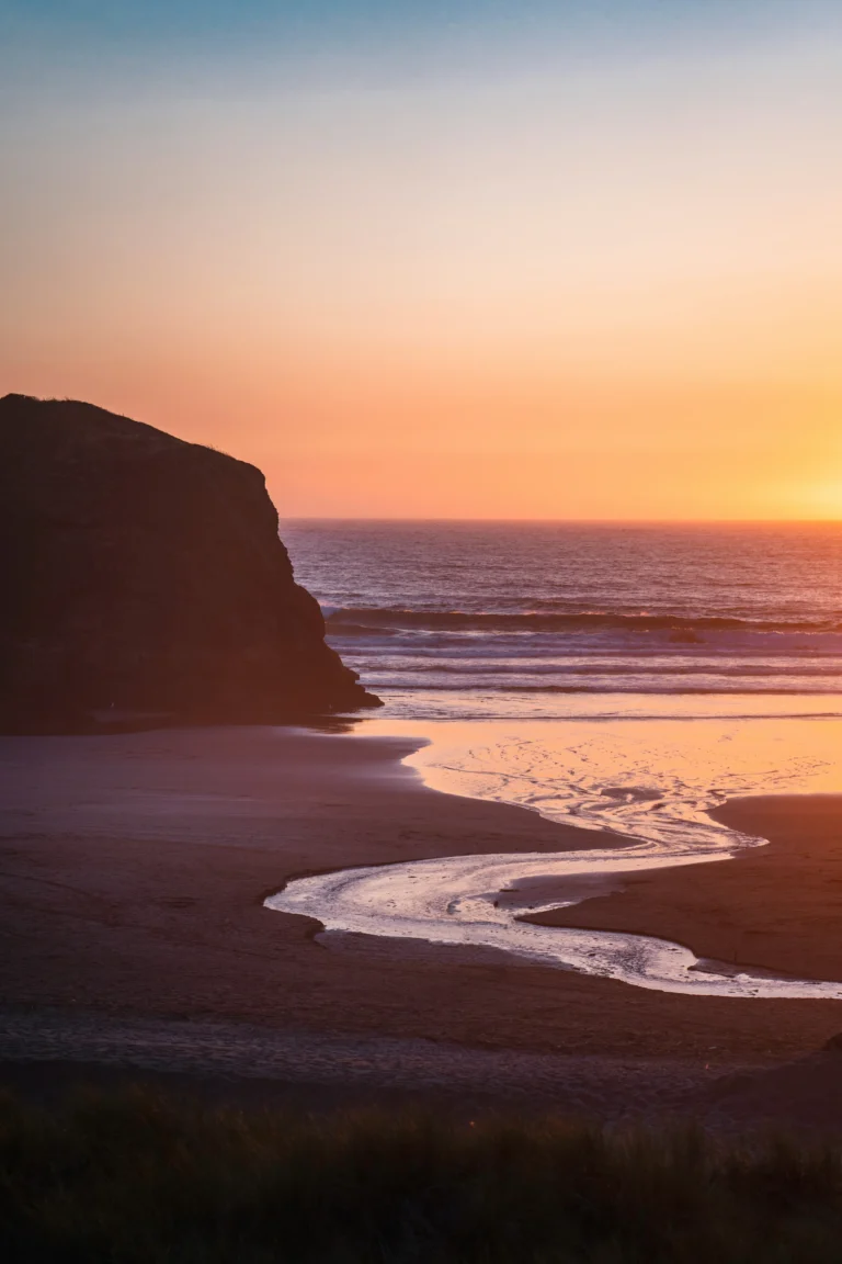 A sunset over a beach with a stream flowing into the ocean, bordered by a large rock formation on the left. The sky displays gradient hues from orange to blue.