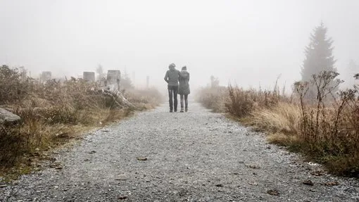A couple walks down a gravel pathway surrounded by fog and dry grass on a misty day.